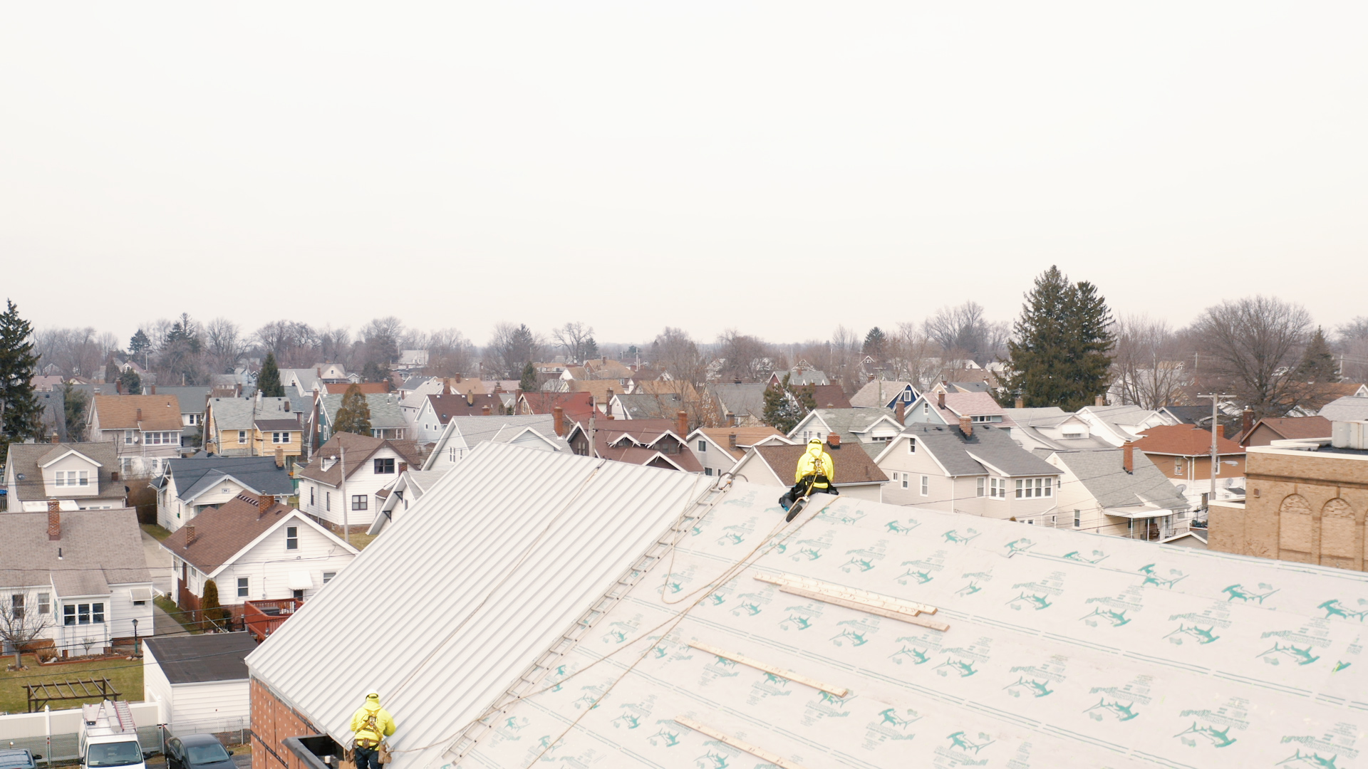 Two roofers equipped with safety harnesses working on installing a new metal roof on a large building.